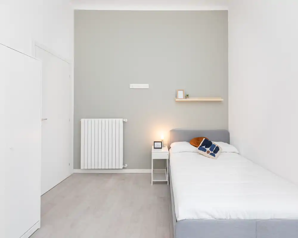 Minimal, bright bedroom with a single bed dressed in white linens, a small bedside table with a lamp, decorative cushions and a floating shelf — ideal for single occupancy or a guest room.