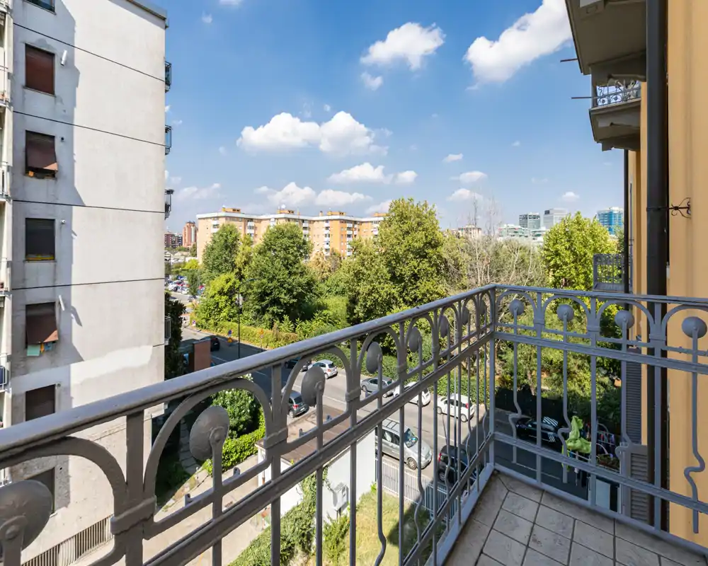 Sunny private balcony with decorative metal railing, tiled floor and pleasant urban view of trees and nearby buildings.