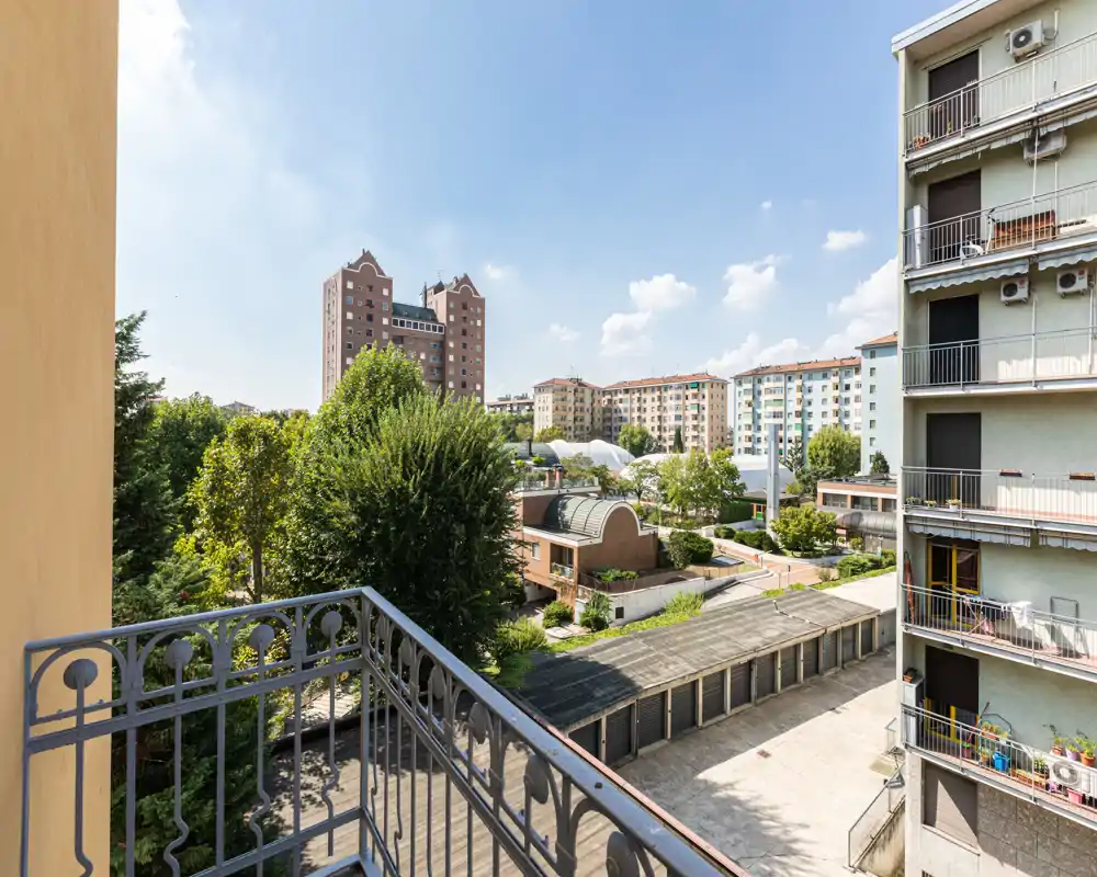 Sunny private balcony with metal railing overlooking trees, neighboring buildings and a courtyard — bright city view ideal for morning coffee.