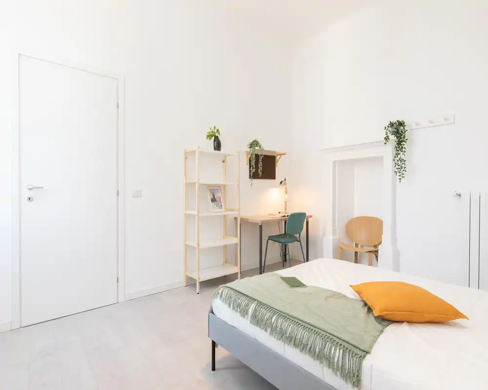 Camera da letto luminosa e minimal con letto ben rifatto, plaid verde e cuscino senape; piccolo angolo studio e scaffalatura rendono l’ambiente accogliente e arioso.