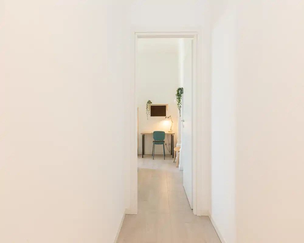 Ingresso-corridoio pulito e minimale che conduce a una stanza luminosa con scrivania; pareti neutre e pavimento in legno chiaro creano un ingresso tranquillo.