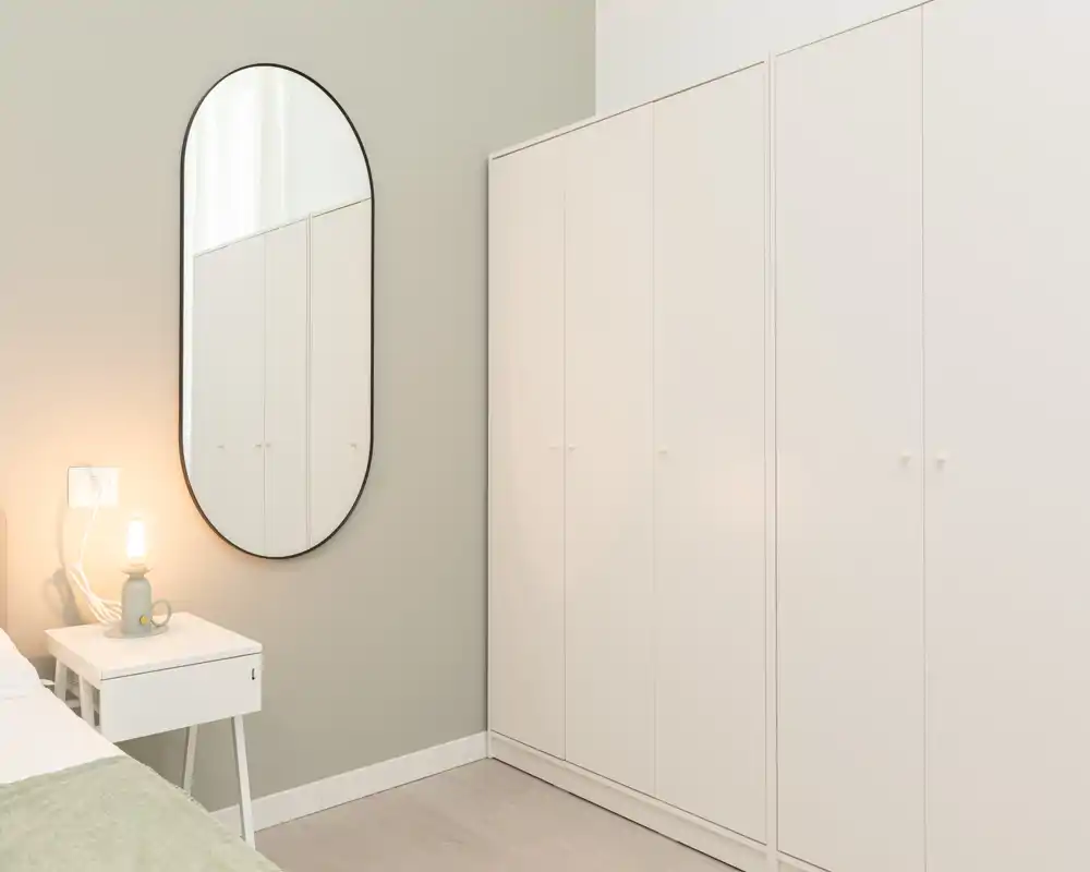 Angolo di camera da letto minimale e luminoso con armadio bianco, comodino e lampada dalla luce soffusa. La palette neutra e lo specchio creano un’atmosfera calma e accogliente, ideale per il riposo.