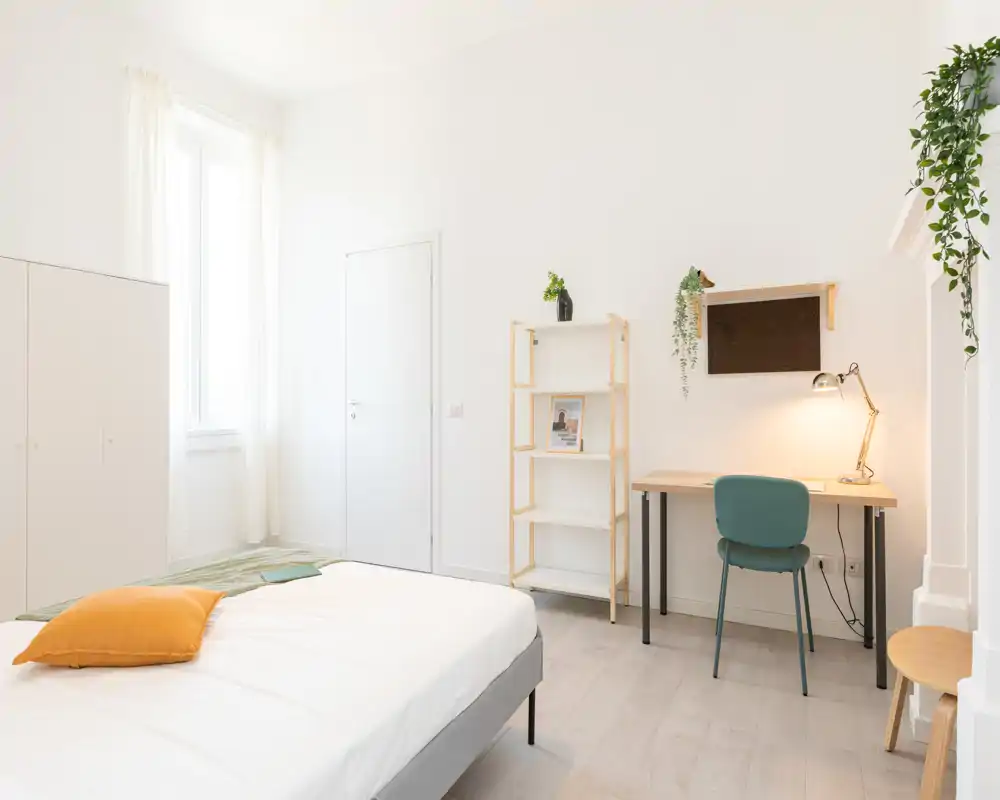Camera da letto luminosa e minimal con letto ben rifatto, scrivania con lampada e mensole; piante che donano un tocco di freschezza.