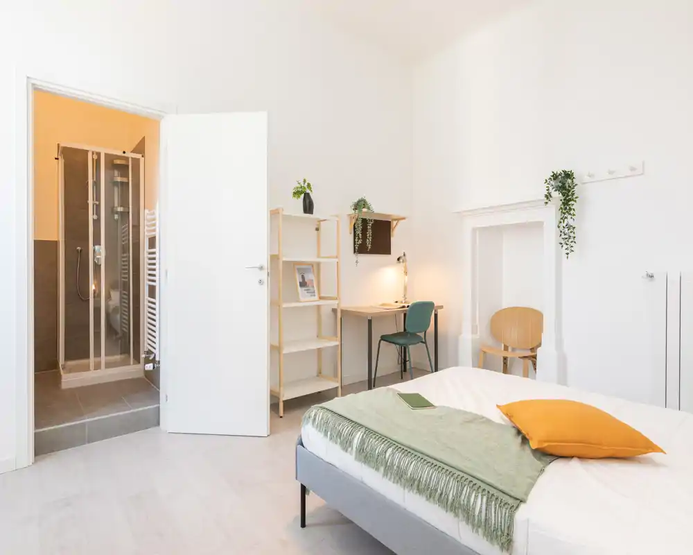 Camera da letto luminosa e minimal con letto matrimoniale in biancheria bianca, un plaid verde e un cuscino arancione, una scrivania con sedia e bagno con doccia visibile dalla porta aperta.