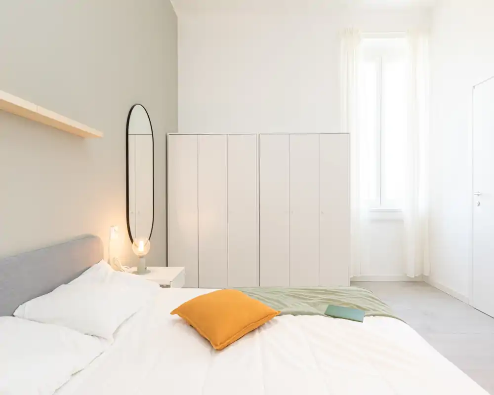 Camera da letto luminosa e minimale con un ampio letto, comodino e armadio; luce naturale soffusa e tocchi caldi la rendono accogliente.