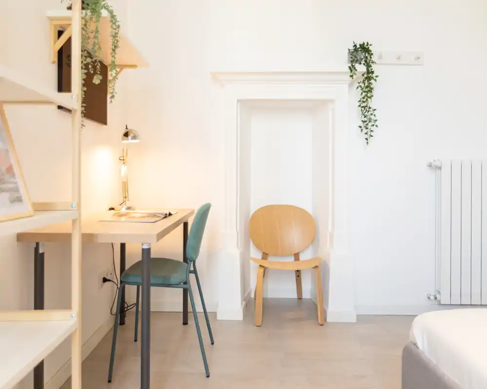 Angolo di camera luminoso e minimal con una scrivania compatta, sedia e una poltroncina in legno; luce soffusa e piante sospese creano un’atmosfera accogliente e rilassante.