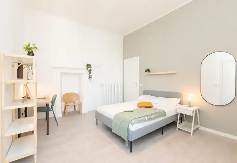 Camera da letto luminosa e moderna con letto matrimoniale ben rifatto, plaid verde, comodino con lampada e grande specchio da parete — stile minimal e arioso perfetto per annunci.