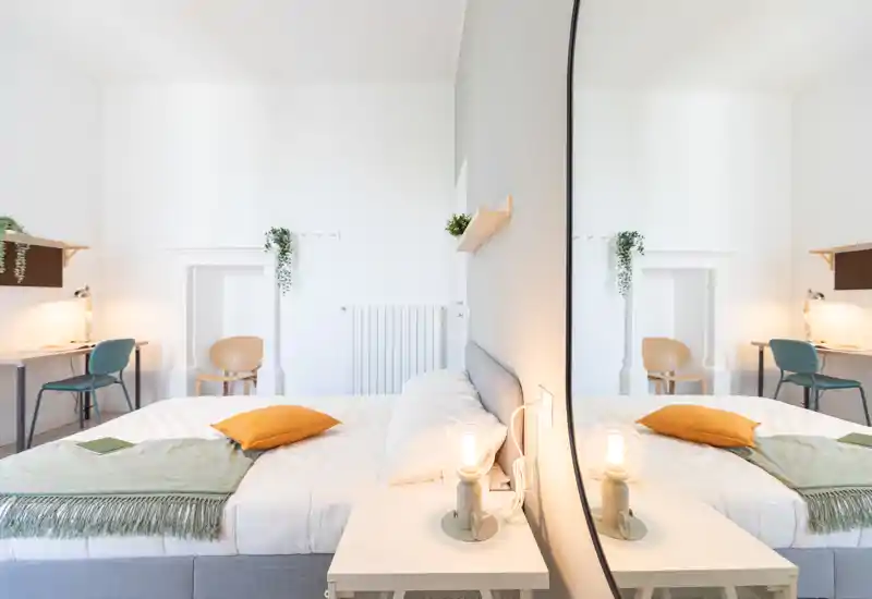 Camera da letto moderna e luminosa con un letto matrimoniale ben rifatto, luce calda sul comodino e una parete specchiata che amplia lo spazio. Due scrivanie con sedie sullo sfondo rendono la stanza funzionale per lavoro e relax.