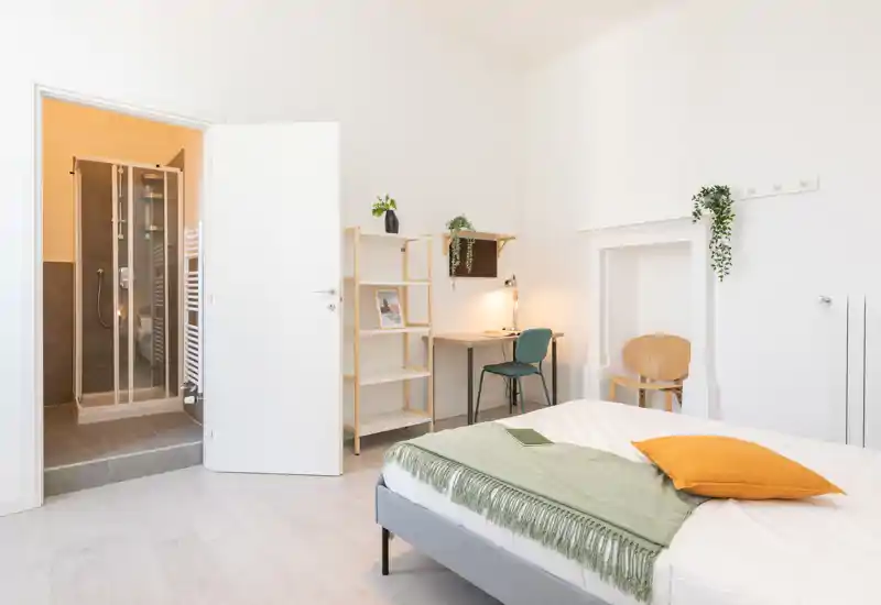Camera da letto luminosa e minimal con letto matrimoniale in biancheria bianca, un plaid verde e un cuscino arancione, una scrivania con sedia e bagno con doccia visibile dalla porta aperta.