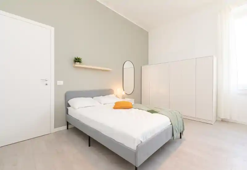 Camera da letto luminosa e minimal con letto matrimoniale completamente visibile, biancheria morbida, armadio e luce naturale da una grande finestra.