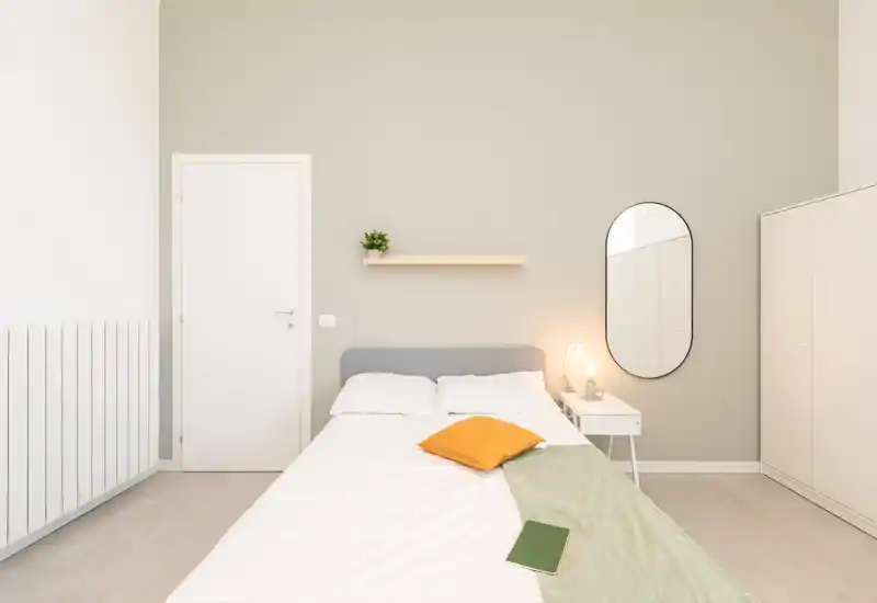 Camera da letto luminosa e minimal con letto matrimoniale, comodino e specchio ovale; toni neutri e tessuti morbidi creano un’atmosfera calma e accogliente.