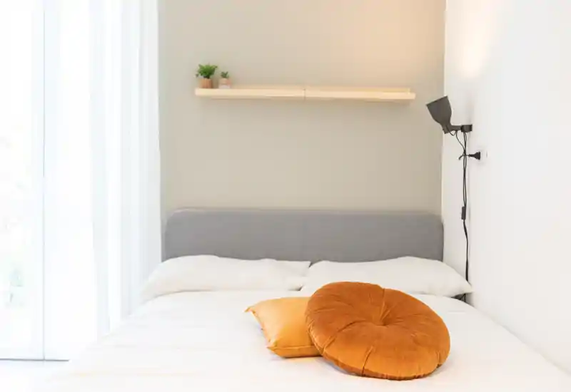 Camera da letto luminosa e minimale con testiera imbottita grigia, biancheria bianca e cuscini decorativi arancioni; luce naturale soffusa dalle tende leggere.
