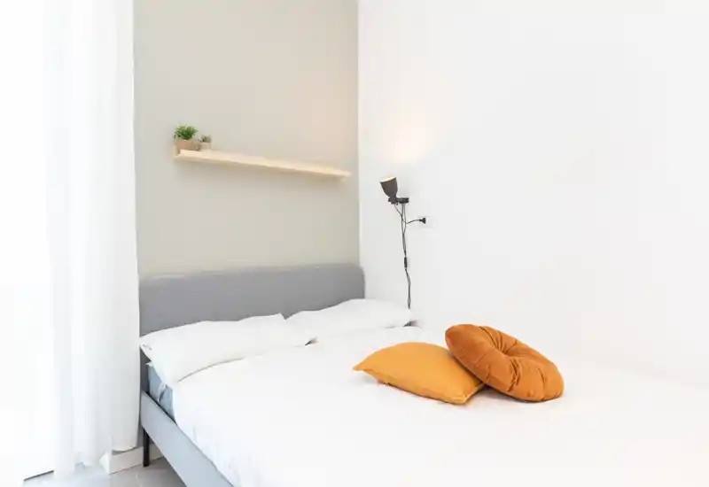 Camera da letto luminosa e minimale con letto imbottito grigio, biancheria bianca e cuscini colorati; la luce naturale dalla finestra e la lampada a muro creano un'atmosfera accogliente.