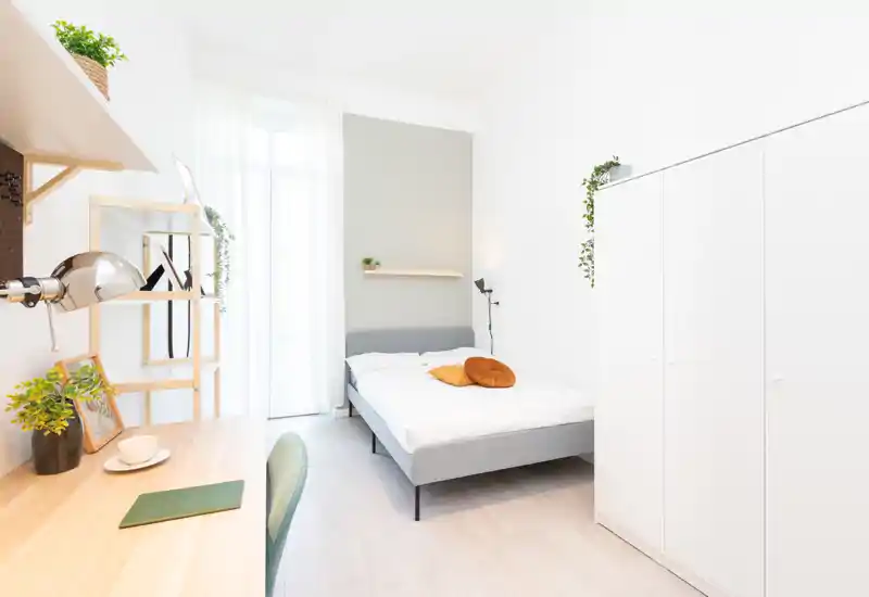 Camera da letto luminosa e minimale con letto matrimoniale, testiera grigia, cuscini d'accento caldi e una scrivania vicino alla finestra. Pareti bianche, ampio armadio e luce naturale che donano un'atmosfera ariosa.