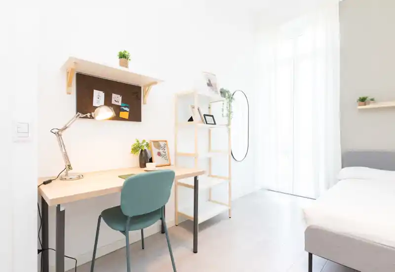 Camera da letto luminosa e minimalista con letto comodo, scrivania in legno e scaffalatura; grande finestra con molta luce naturale.