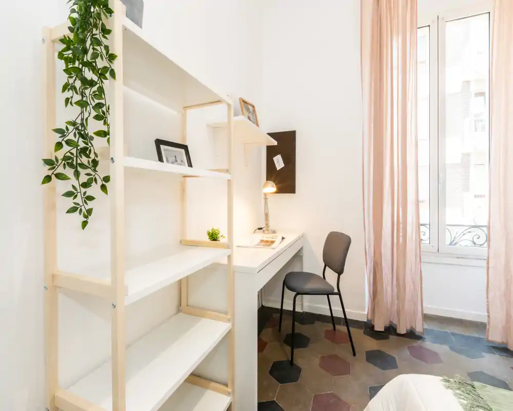 Angolo camera da letto luminoso e arredato con scrivania, sedia e scaffale; tende leggere e piante decorative creano un'atmosfera accogliente, perfetta per un affitto breve.