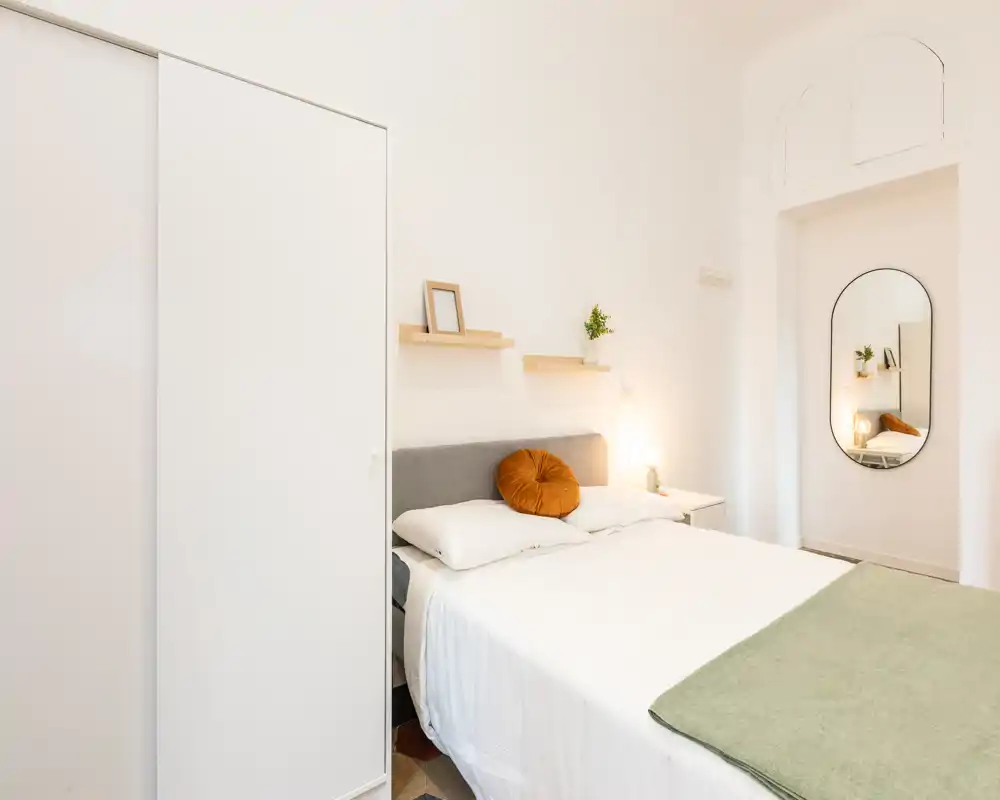 Camera da letto luminosa e minimal con un comodo letto matrimoniale, cuscino decorativo caldo e uno specchio a figura intera che riflette una luce soffusa. Armadio pratico e mensole semplici creano uno spazio calmo e di stile, ideale per soggiorni riposanti.