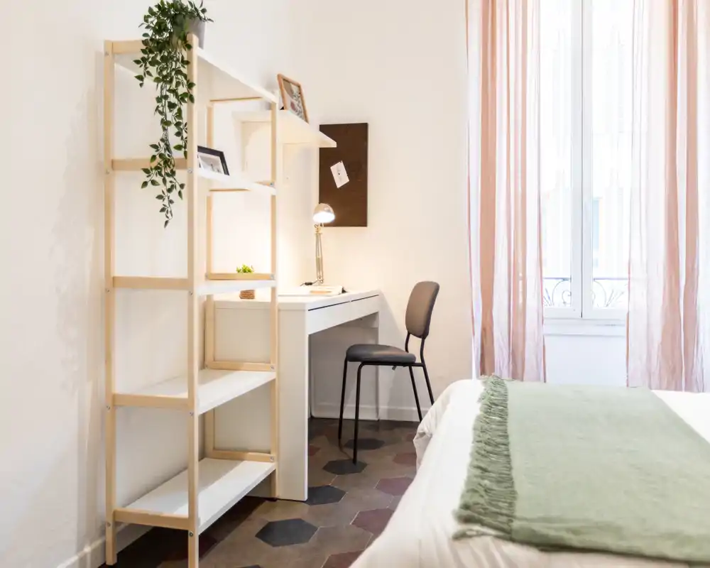 Camera da letto luminosa e minimal con letto in primo piano, scrivania ordinata e sedia accanto alla finestra, e scaffalatura con piante e oggetti decorativi per un’atmosfera moderna e accogliente.