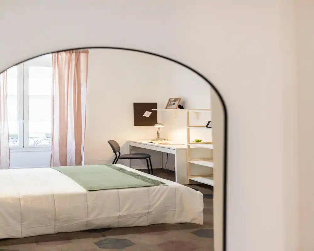 Camera da letto luminosa e curata con letto matrimoniale ben rifatto, plaid verde, scrivania con lampada e scaffalatura, illuminata da luce naturale attraverso tende chiare.