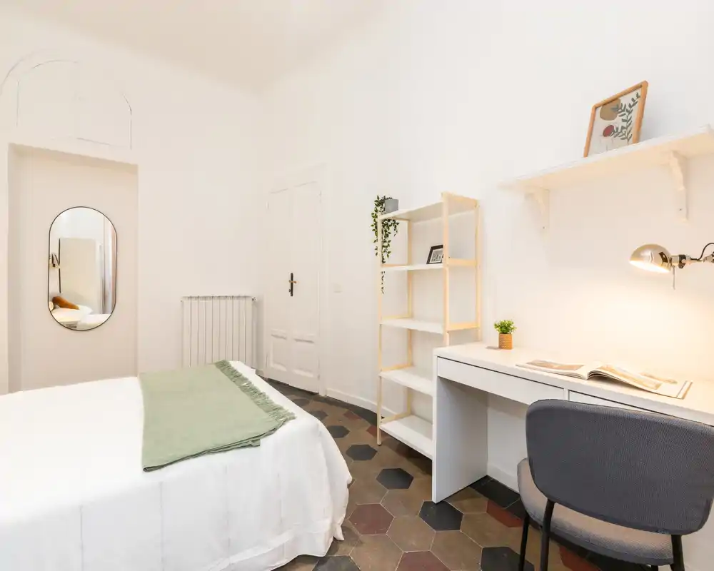 Camera da letto luminosa e minimal con comodo letto matrimoniale, scrivania ordinata e scaffalatura — pareti bianche e pavimento a piastrelle donano un'atmosfera moderna e rilassante.