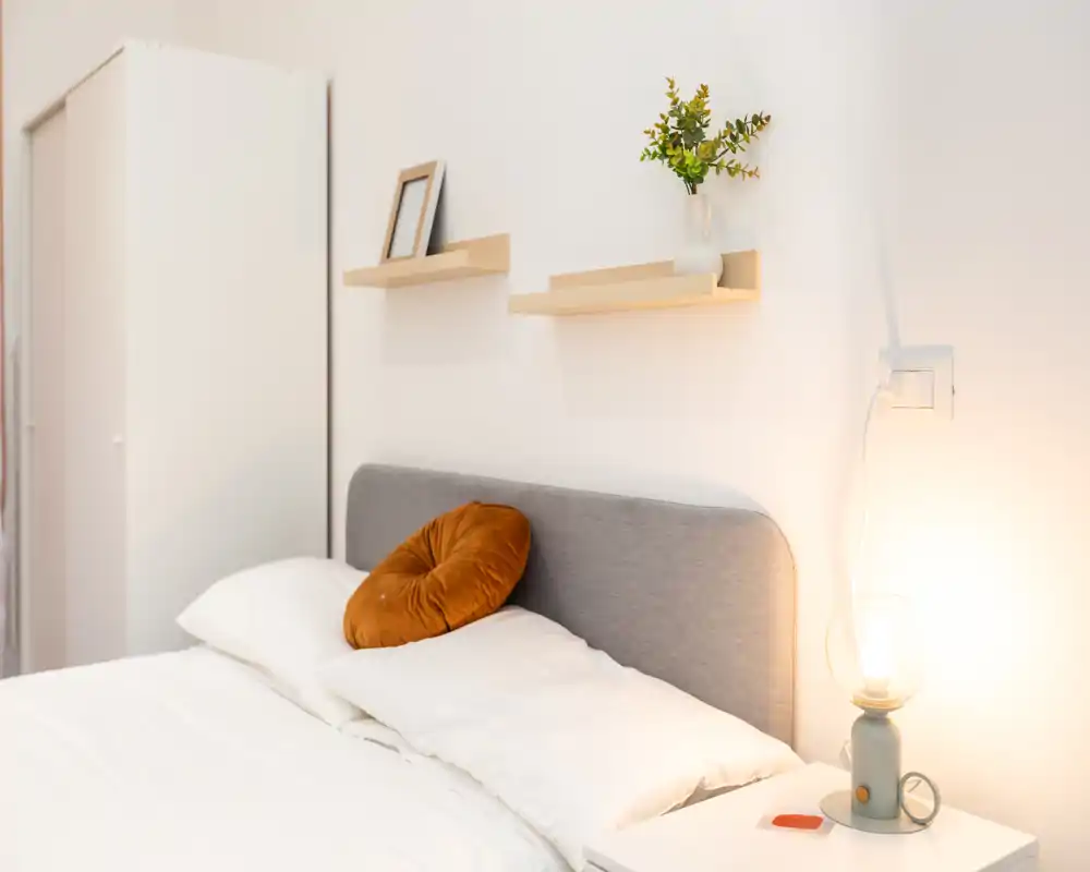Camera da letto luminosa e minimal con testiera imbottita grigia, biancheria bianca e un cuscino decorativo; mensole a muro e una lampada da comodino creano un’atmosfera accogliente.