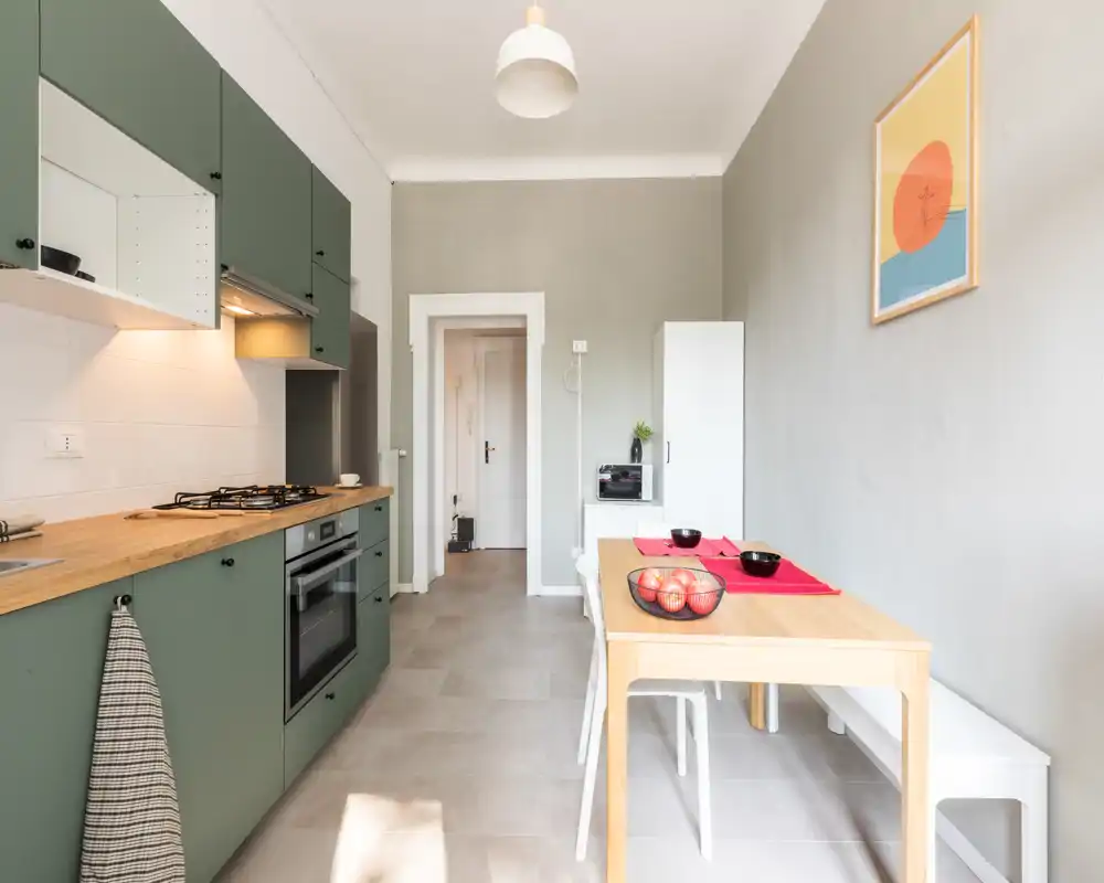 Cucina moderna e compatta con pensili verdi, piano in legno, forno incassato e piccolo tavolo da pranzo — luminosa e ben arredata per una cucina funzionale.