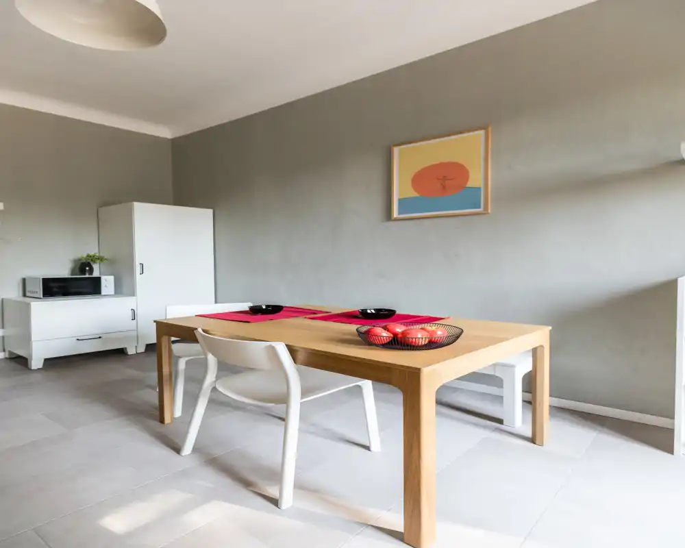 Zona pranzo minimal e moderna con tavolo in legno chiaro, sedie bianche essenziali e ciotole decorative; ambiente luminoso e pulito perfetto per le foto dell'annuncio.