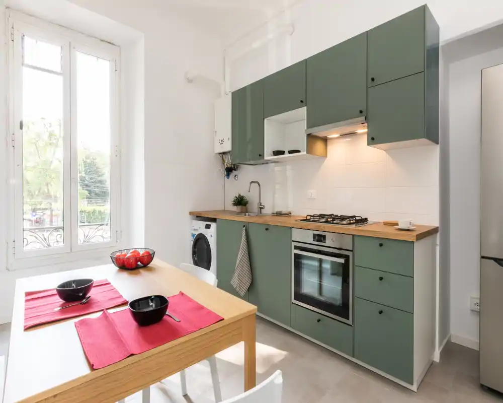 Cucina moderna e luminosa con pensili verdi, piano in legno e forno integrato; tavolo da pranzo vicino a una grande finestra per una colazione accogliente.