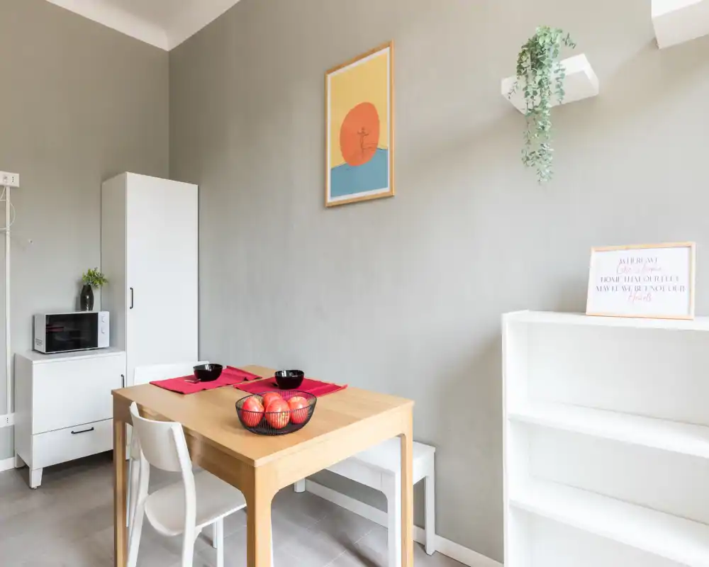 Piccolo angolo pranzo luminoso con tavolo in legno per due, scaffalatura minimal e mobile con microonde — pulito e accogliente per pasti informali.