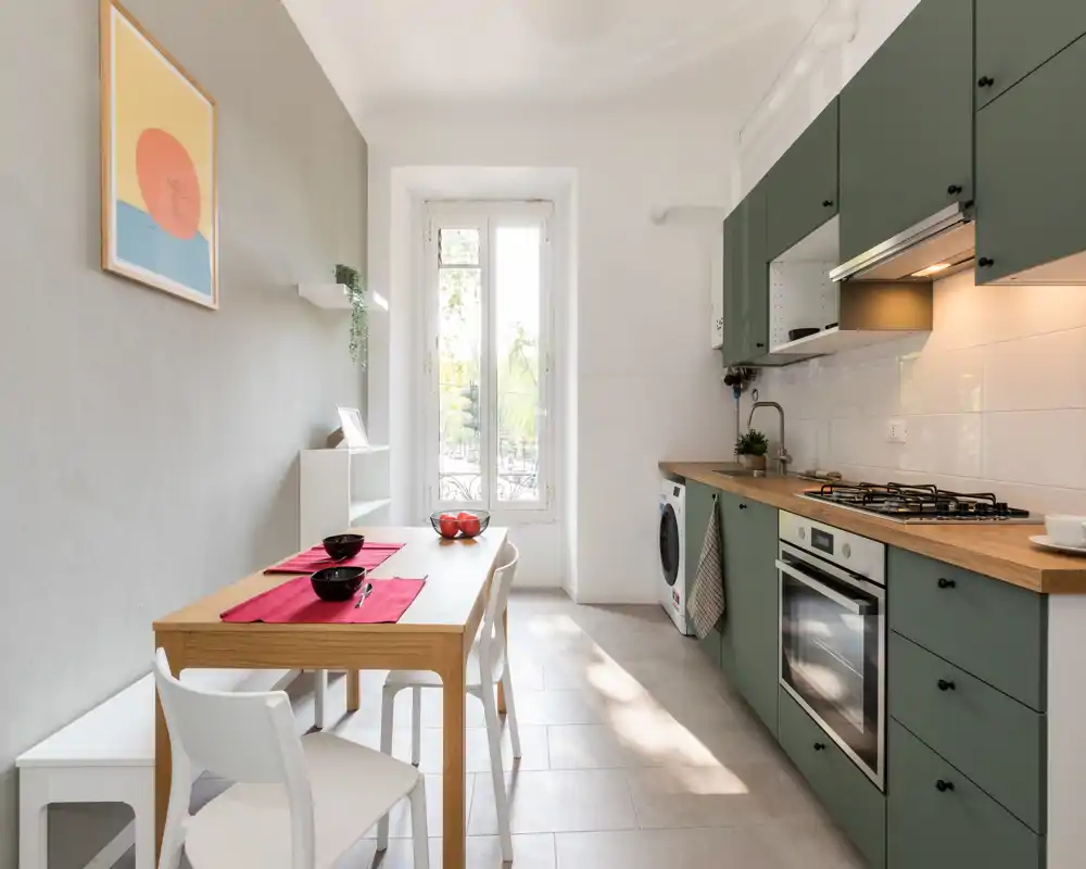 Cucina moderna a corridoio luminosa con pensili verdi, piani in legno, forno incassato e piano cottura a gas, tavolo per due e molta luce naturale da una finestra alta.