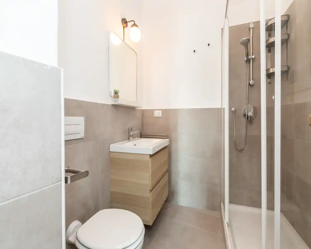 Bagno moderno e compatto con doccia angolare, mobile lavabo sospeso e WC. Piastrelle neutre e illuminazione soffusa creano uno spazio pulito e funzionale.
