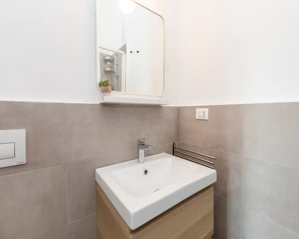 Compatto bagno moderno in angolo con lavabo bianco su mobile in legno chiaro, piastrelle a parete, specchio e piccolo ripiano — luminoso, pulito e pronto all'uso.