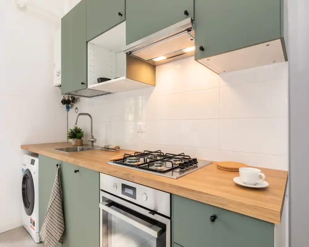 Angolo cottura moderno e luminoso con pensili verdi, piano in legno, piano cottura a gas e forno incassato. Layout compatto e ordinato con lavatrice e illuminazione sotto i pensili.
