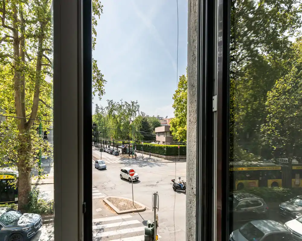 Vista sulla strada da una finestra che si affaccia su un incrocio soleggiato incorniciato da alberi e auto in transito; panorama urbano piacevole con buona visibilità della strada e degli edifici vicini.