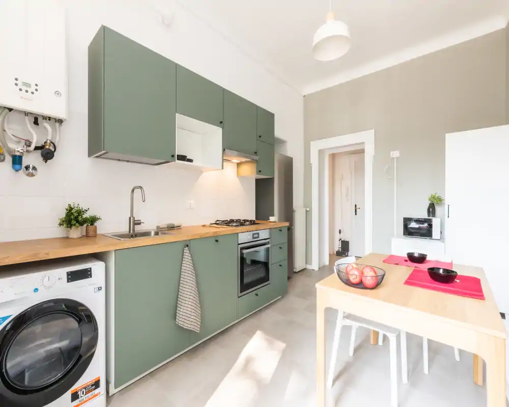 Cucina moderna e luminosa con mobili verdi, piano in legno, forno incasso e piccolo tavolo da pranzo — spazio funzionale e curato, perfetto per annunci di appartamento.