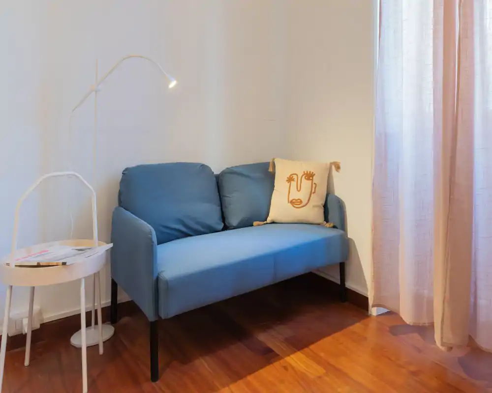 Compact living area with a two-seater blue sofa, a decorative cushion and warm wooden flooring creating a cozy corner with soft natural light from the curtained window.