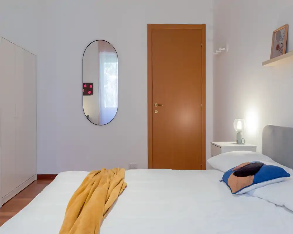 Minimal, bright bedroom with a neatly made bed, bedside table with lamp, mirror on the wall and a wardrobe — clean modern styling ideal for rental photos.