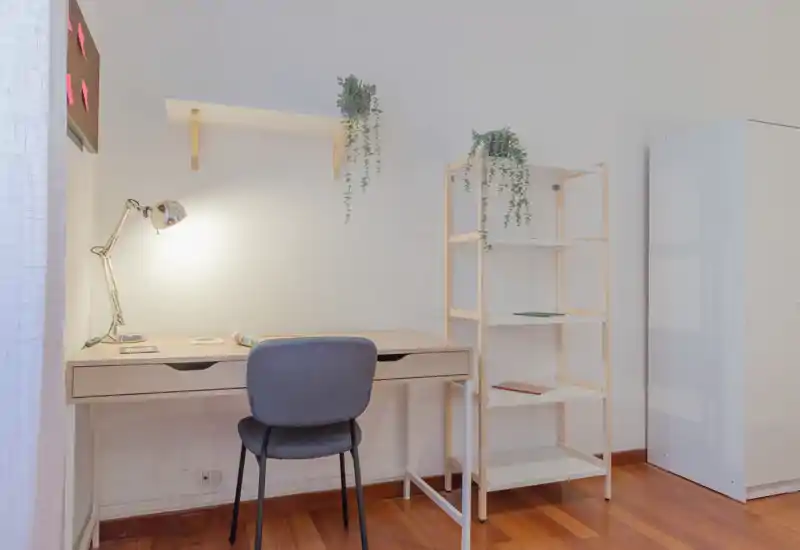 Angolo studio ordinato con scrivania in legno, lampada da tavolo, sedia confortevole e scaffalatura aperta; minimal, luminoso e adatto per studiare o lavorare da remoto.
