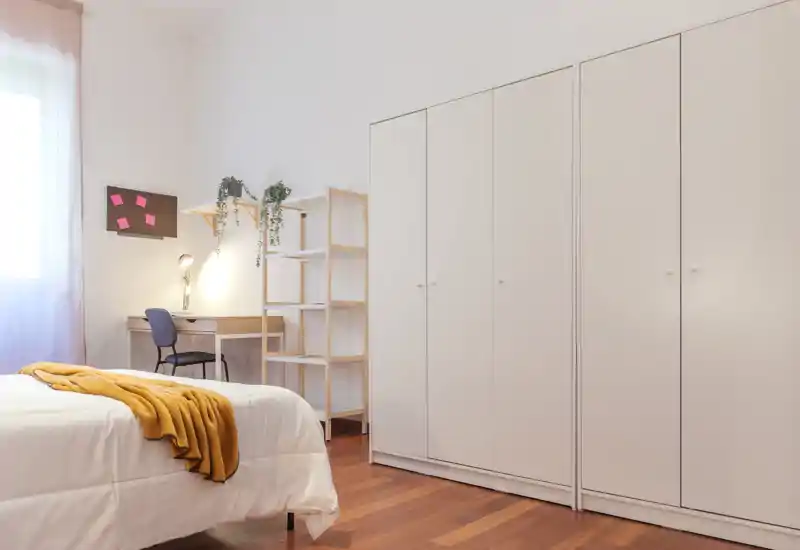 Bright, minimal bedroom with a comfortable bed, a work desk by the window and large built-in wardrobes. Natural light and warm wooden floors create a welcoming atmosphere.