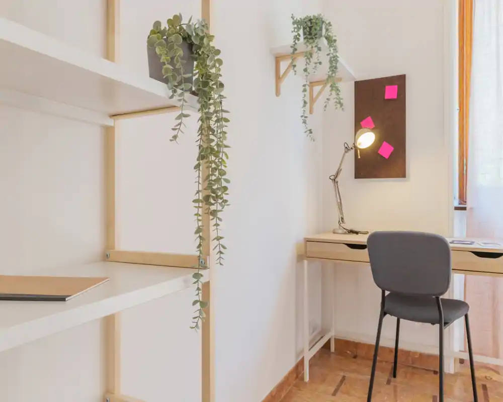 Zona de estar luminosa y minimalista con un rincón de trabajo ordenado: escritorio de madera con lámpara y silla junto a la ventana, estanterías abiertas y plantas colgantes que aportan un toque fresco y acogedor.