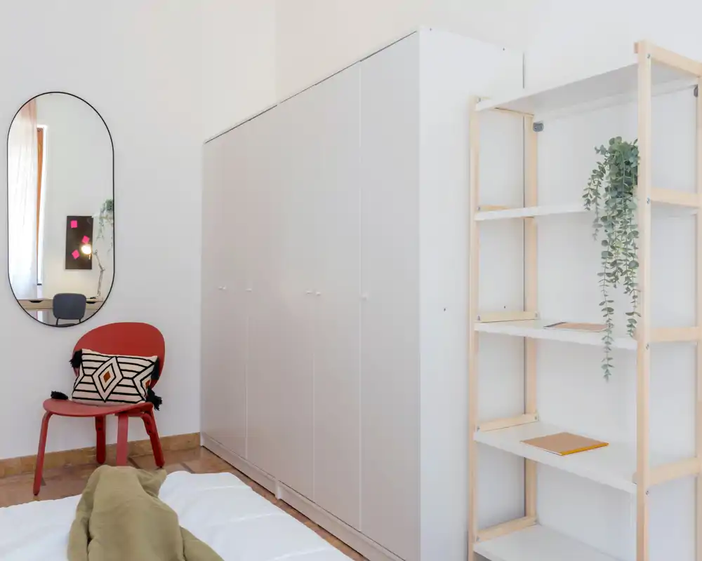 Rincón de dormitorio luminoso y minimalista con cama en primer plano, gran armario blanco, estantería abierta y una silla decorativa con cojín que aporta calidez.