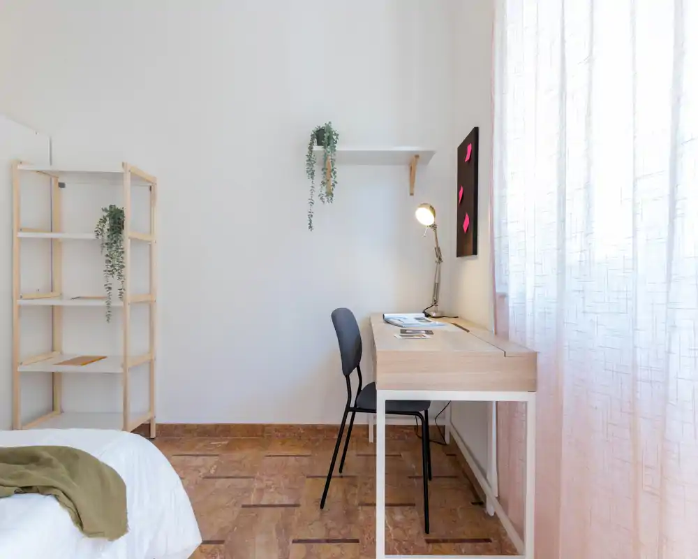 Rincón de dormitorio luminoso y minimalista con el borde de la cama visible, un escritorio junto a la ventana, estantería y cortinas ligeras — ideal para una persona o espacio de estudio.