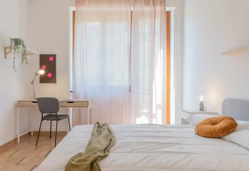 Camera da letto luminosa e minimal: un ampio letto in primo piano, luce naturale morbida attraverso tende leggere, un comodino e una pratica scrivania vicino alla finestra.