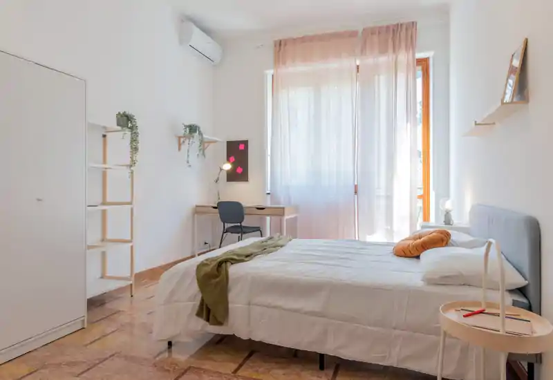 Camera da letto luminosa e ben allestita con un letto matrimoniale, tessili morbidi e una scrivania vicino alla finestra soleggiata. Pavimento caldo e mensole minimal per un'atmosfera moderna e tranquilla.