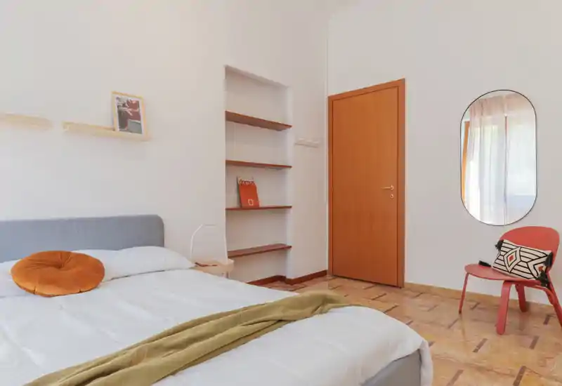 Camera da letto moderna e luminosa con comodo letto matrimoniale, tessili caldi e mensole minimal — perfetta per un soggiorno accogliente.