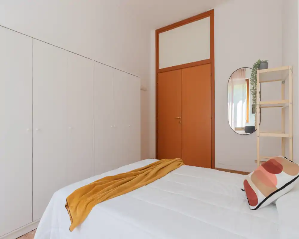 Bright, minimalist bedroom with a large white bed dressed with a mustard throw and patterned cushion, a tall white wardrobe and a wooden door; neat, well-lit and ready for guests.