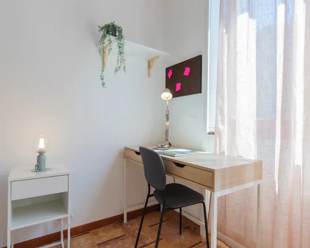 Bright, minimal bedroom corner arranged as a small workspace with a wooden desk, chair, lamp and soft natural light from a curtained window.