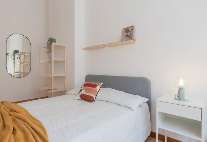 Dormitorio luminoso y minimalista con una cama bien hecha, cojín decorativo y una cálida manta. Estanterías sencillas, espejo y mesita de noche con lámpara crean un espacio acogedor y bien presentado, ideal para anuncios.