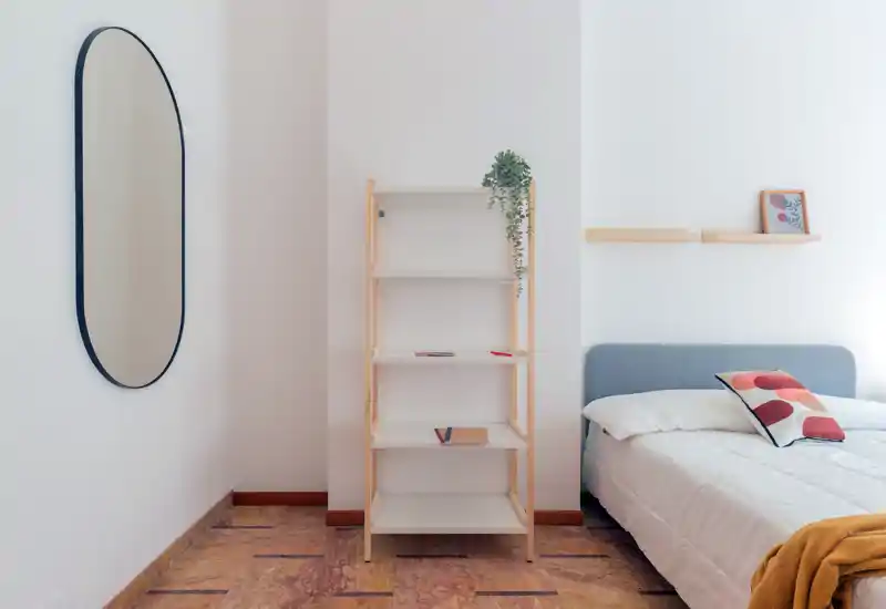 Rincón de dormitorio minimalista y luminoso con cama bien hecha, cojines decorativos y estantería de madera; líneas limpias y luz natural suave que transmiten calma y modernidad.