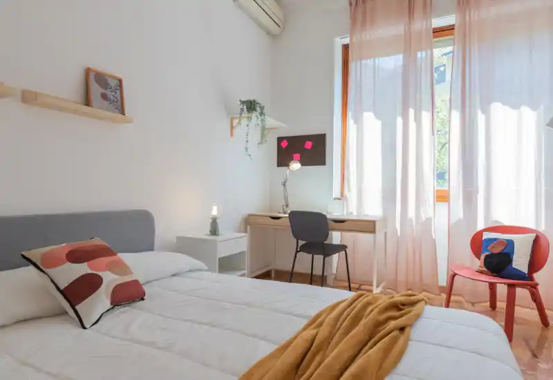 Dormitorio moderno y luminoso con cama bien hecha, textiles suaves y un pequeño espacio de trabajo junto a la ventana — ideal para estancias cómodas y teletrabajo.